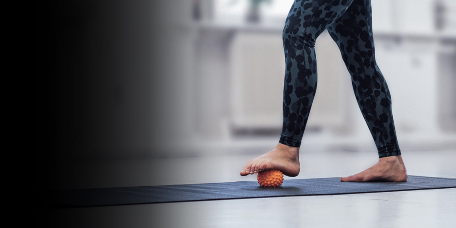 Female in yoga pants and bare feet using an orange foot massage ball on a yoga mat. Image fades from black to transparent from left to right