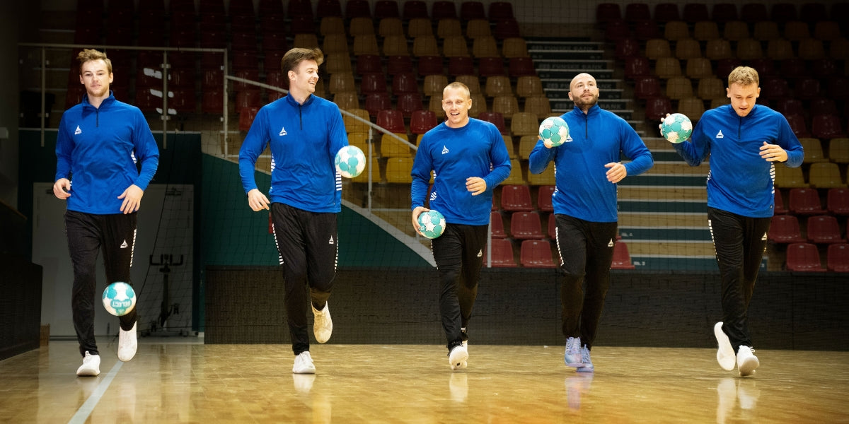 Handball players wearing blue jackets and black pants and dribbling balls on a court