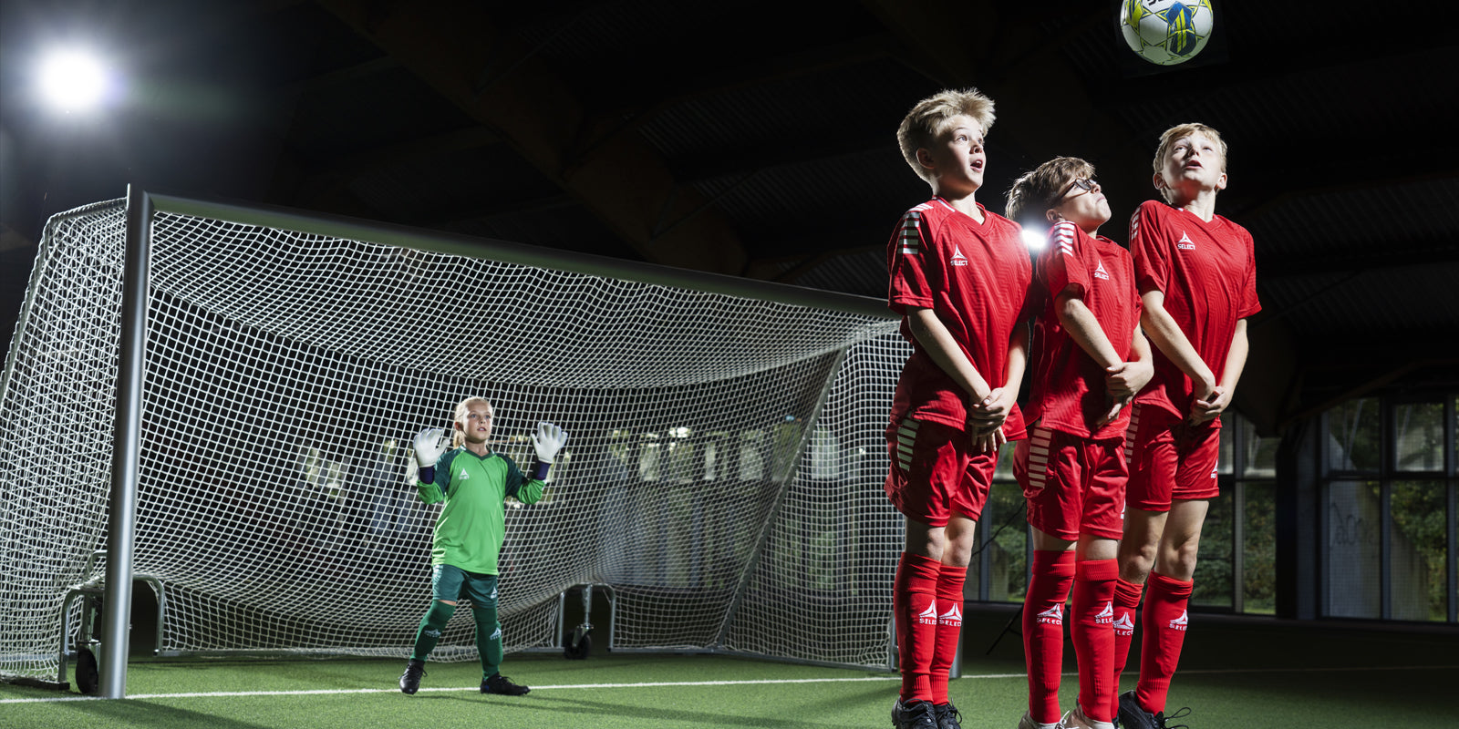 Three soccer players in red uniforms with a ball going over their head toward a goal with a goalkeeper wearing green