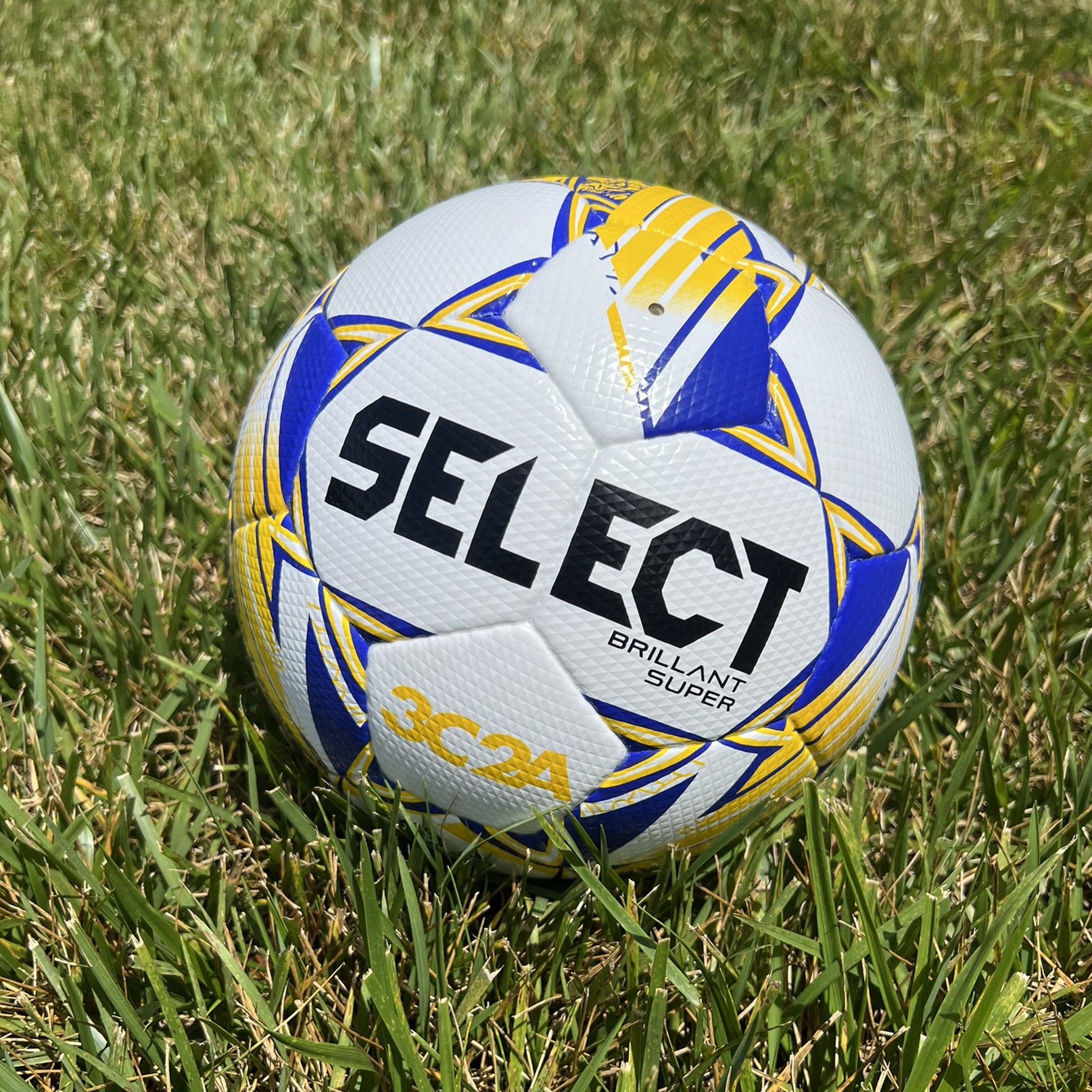 White soccer ball with blue and yellow accents and 'Select' branding on a grass field. Used by the 3C2A