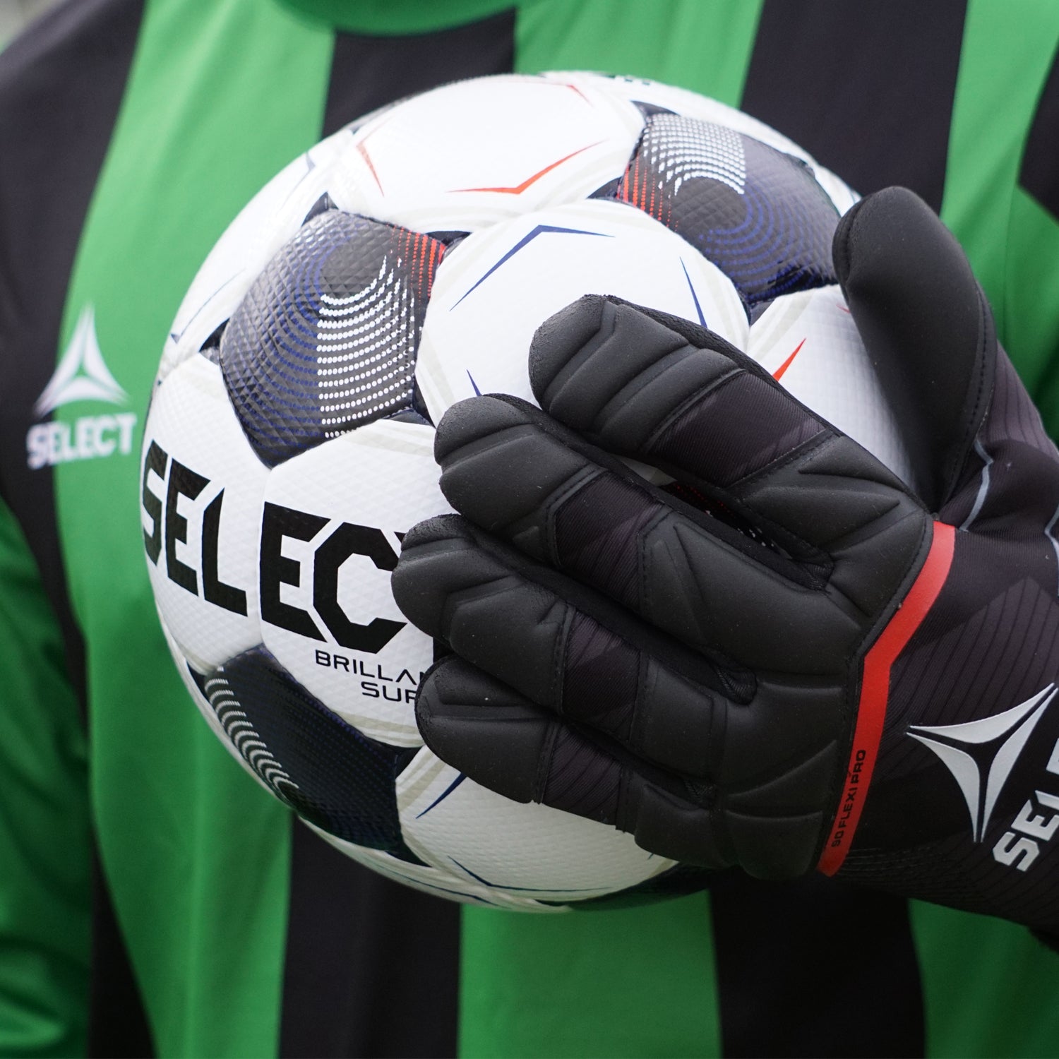 Soccer goalkeeper in a black and green jersey wearing black and red goalkeeper gloves holding a white soccer ball with black, blue, and red coloring branded with 'Select'.