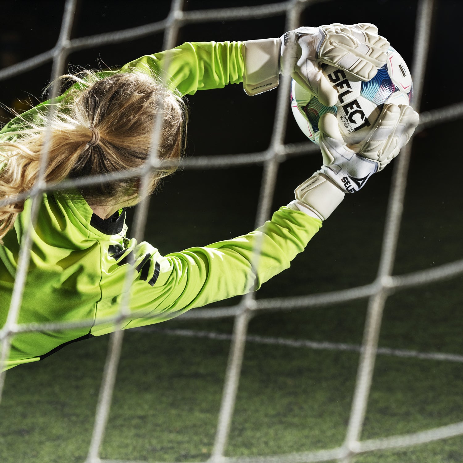 Goalkeeper in soccer net catching a soccer ball wearing white goalkeeper gloves and yellow long sleeve shirt