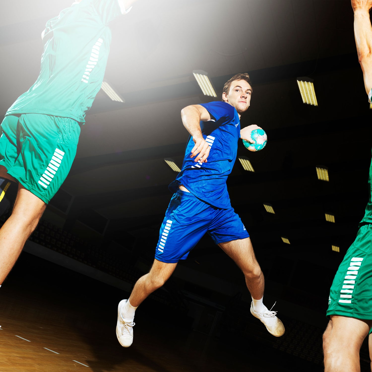 Handball players in a gym wearing blue or green uniforms in action throwing a teal handball