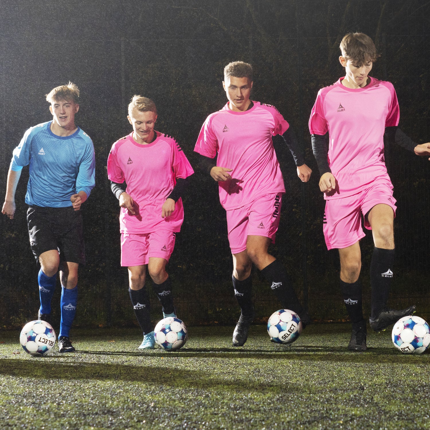 Male soccer players wearing pink uniforms and one player in a blue goalkeeper uniform dribbling soccer balls. Clothing and balls branded with Select logo