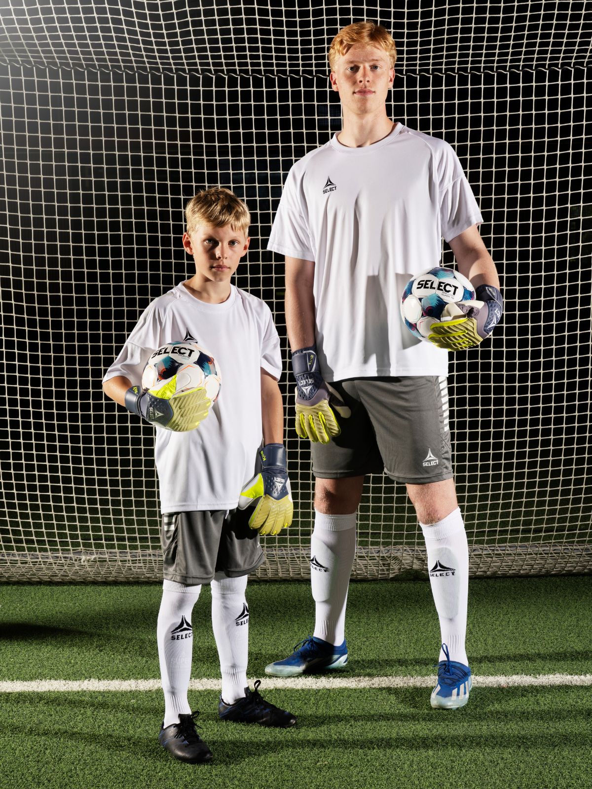 Two young soccer players standing in front of a goal, wearing matching sports outfits with gloves and cleats.