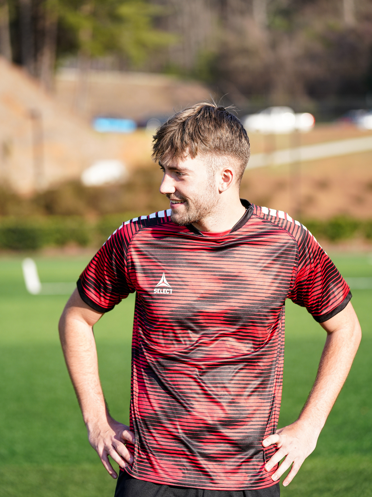Soccer player wearing a red and black wavy jersey with a white Select logo on an outdoor soccer field