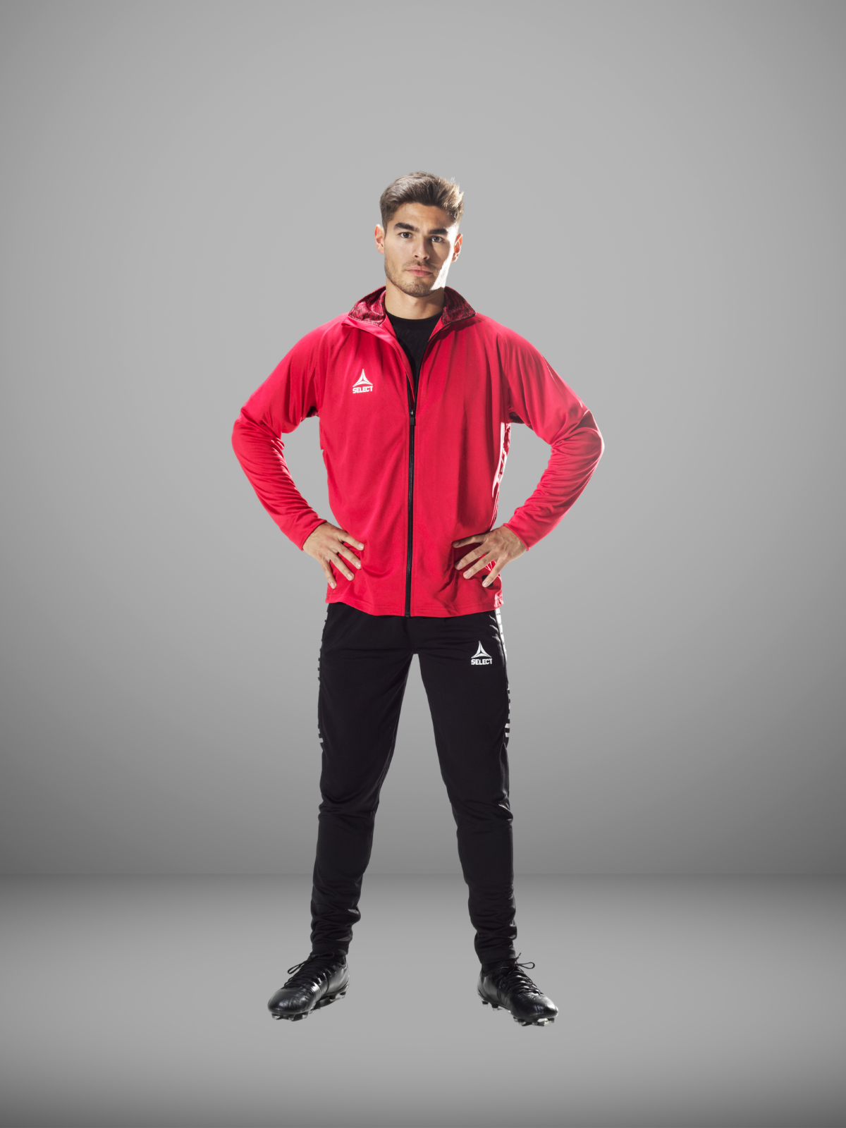 Soccer player in a red full zip jacket, black pants and black cleats on a grey background