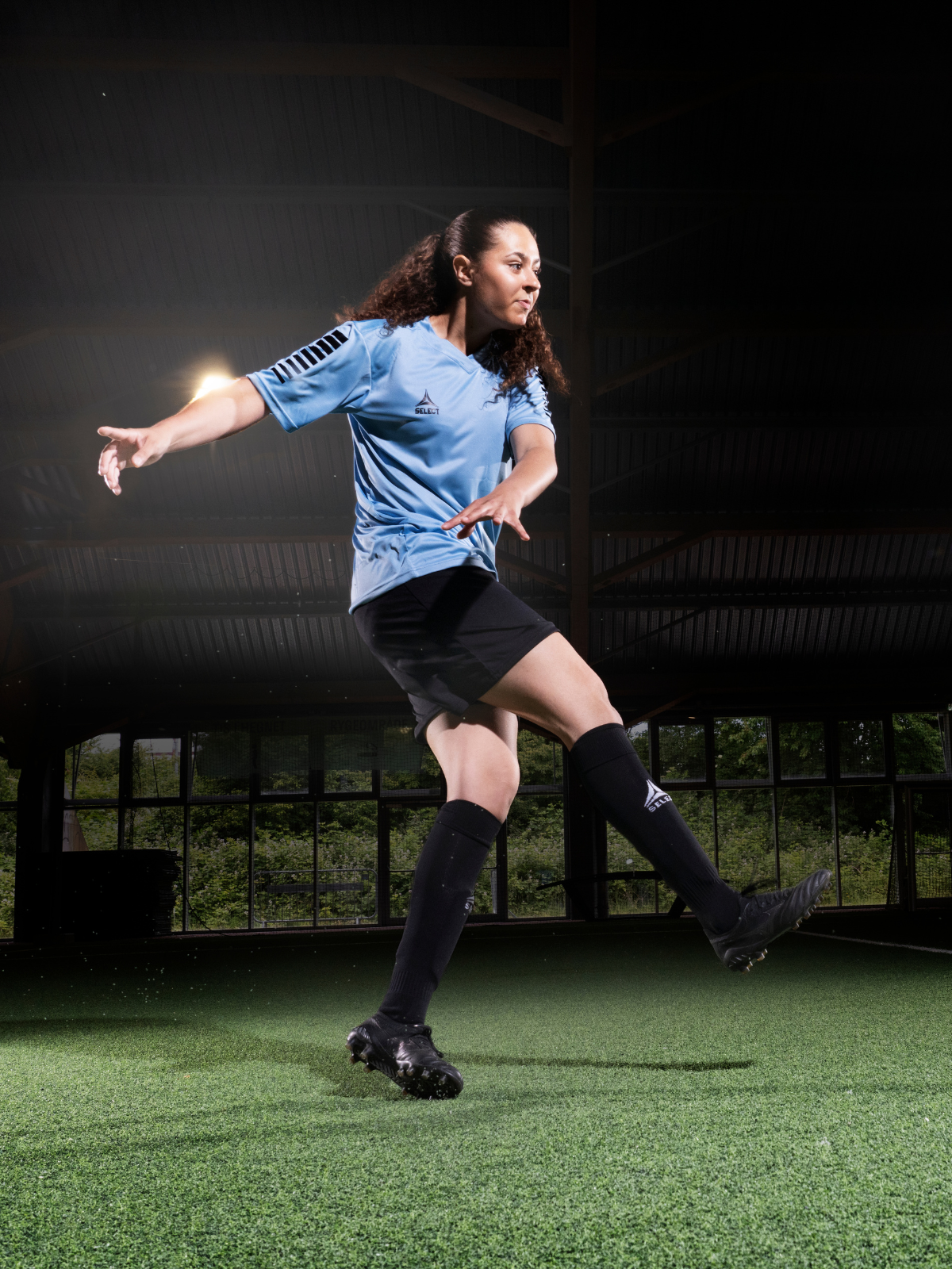 Soccer player wearing a light blue jersey, black shorts, and black socks in a kicking motion on an indoor soccer field with a dark background