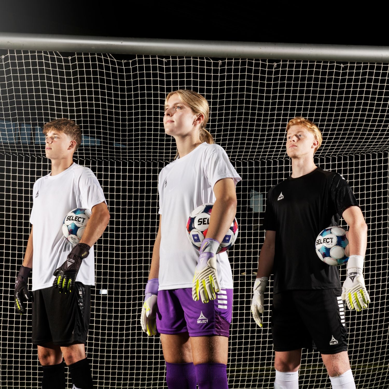 Three soccer goalkeeper standing in front of a net, each holding a soccer ball.